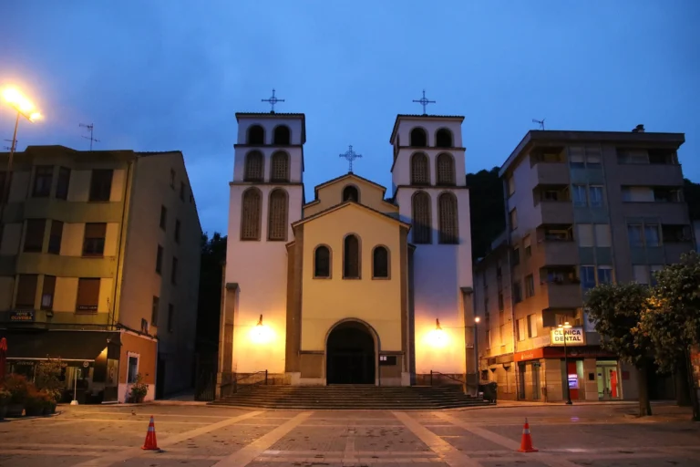 Iglesia de dos torres en el centro de Pola de Lena al anochecer, con la plaza iluminada delante