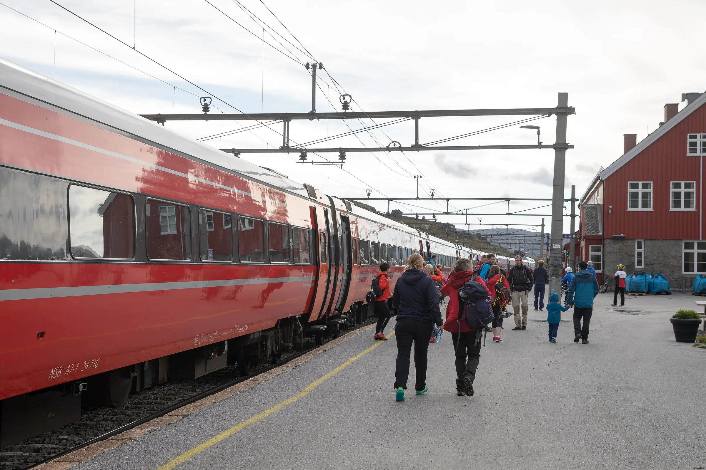 Tren rojo de Vy parado en el andén de la estación de Finse, con viajeros y el edificio rojo de la estación al fondo
